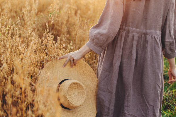 Stylish woman with straw hat walking in oat field in sunset light, cropped view. Young female in rustic linen dress relaxing in evening summer countryside. Rural slow life