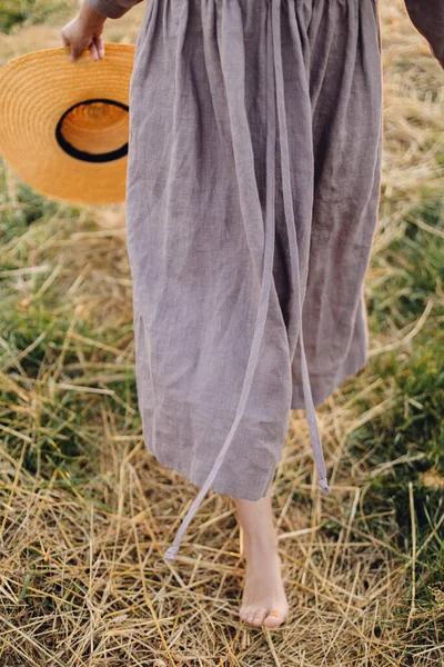 日没の光の中で麦わら畑を裸足で歩いて手に帽子を持つ女性は 閉じます 農村部のスローライフ 落ち着いた雰囲気の瞬間 若いです女性で素朴なリネンドレスで夏の田舎でリラックス — ストック写真