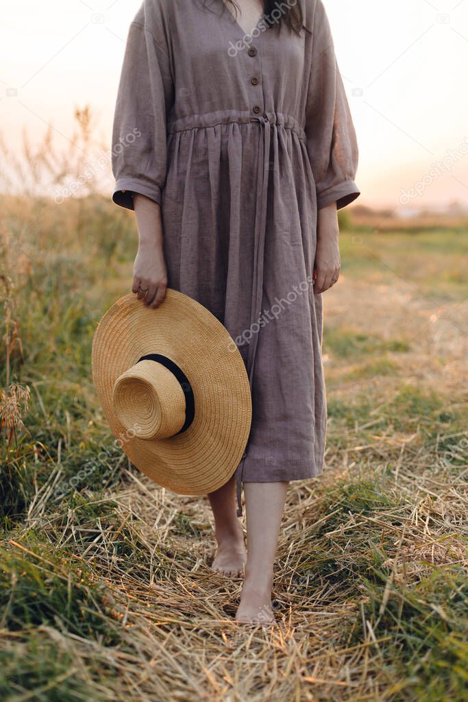 Woman with hat in hand walking barefoot on straw field in sunset light, cropped view. Rural slow life. Atmospheric tranquil moment. Young female in rustic linen dress relaxing in summer countryside