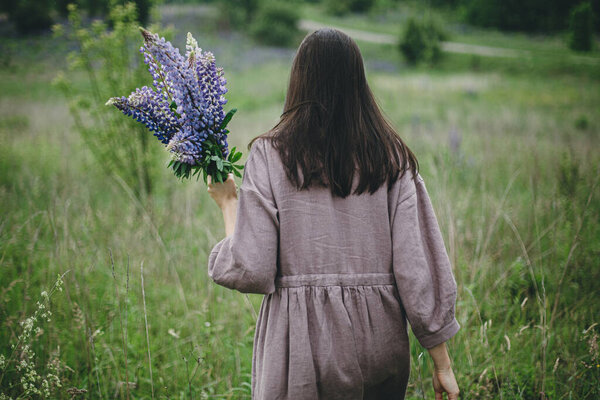 Stylish woman in rustic dress gathering lupine bouquet in meadow, atmospheric moment. Cottagecore aesthetics. Young female in linen dress holding wildflowers in summer countryside, rural slow life