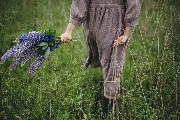 Close up of woman in rustic dress holding lupine bouquet in meadow. Cottagecore aesthetics. Young female in linen dress walking with wildflowers in atmospheric summer countryside, rural slow life