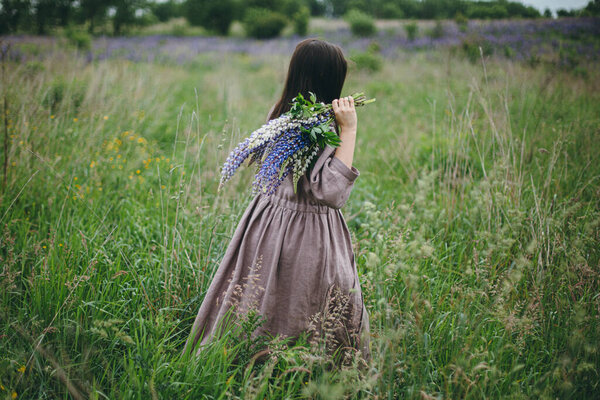 Stylish woman in rustic dress walking among lupine meadow, atmospheric image. Young female in linen dress gathering flowers in summer countryside, rural slow life. Cottagecore aesthetics
