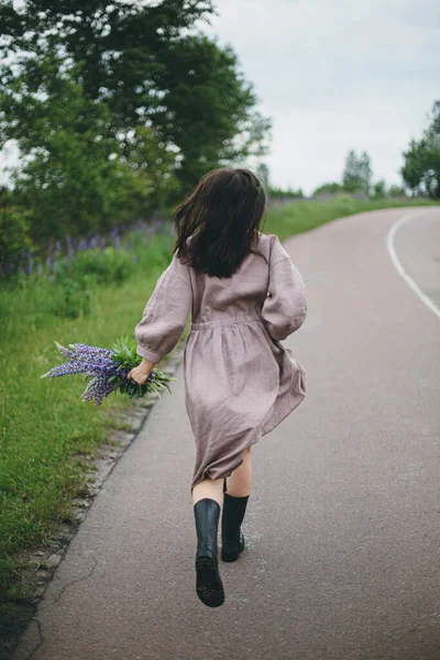 夏天乡村 穿着乡村风情服装 头戴紫丁香花束跑步的时髦女人 土工布美学 乡村道路上的雨后 身穿亚麻布衣服的年轻女子穿着野花休闲装 — 图库照片