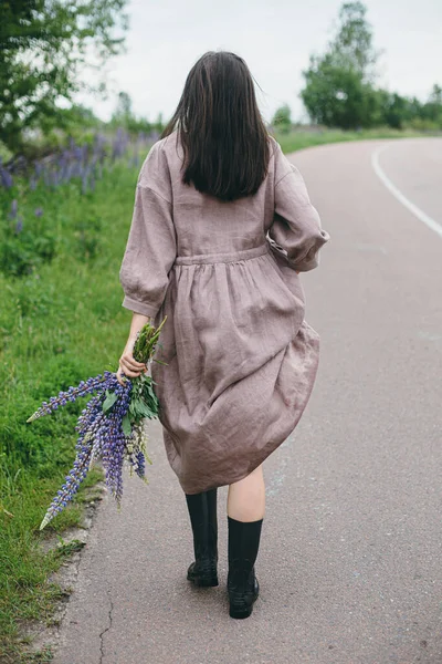 夏天的乡间 穿着乡村风情服装 头戴紫丁香花束的时髦女人在散步 土工布美学 穿着亚麻布衣服的年轻女子 雨后抱着野花在乡间路上 生活节奏缓慢 — 图库照片
