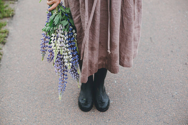 Close up of woman in rustic dress and gumboots holding lupine bouquet in summer countryside. Cottagecore aesthetics. Young female with wildflowers after rain on rural road.Moody image