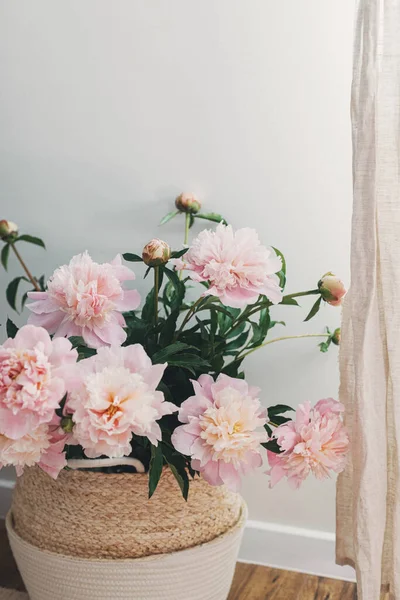 Beautiful peonies bouquet in basket in modern boho room. Gentle pink peony flowers on rustic background, moody image. Modern bohemian decor, stylish comfy interior details