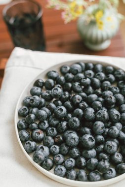 Modern seramik tabakta taze yaban mersini ve kır evinde kır çiçeği buketi. Kırsal kesimde yaz mevsimi. Sağlıklı gıda estetiği. Yaz meyveleri