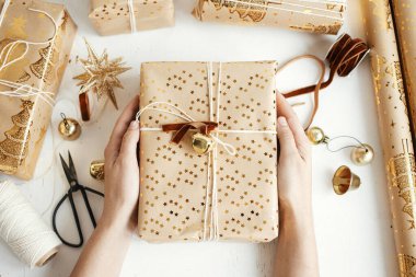 Merry Christmas! Hands holding stylish golden christmas gifts flat lay. Person preparing modern christmas gift boxes, festive paper, golden ornaments and thread composition on white wood