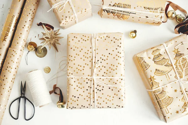 Stylish christmas gift flat lay. Modern wrapped christmas gift boxes, festive paper, golden ornaments and thread composition on white wooden background. Merry Christmas!