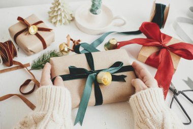 Merry Christmas! Hands wrapping stylish christmas gift. Person preparing  christmas present with green ribbon and ornaments composition on white wood. Aesthetic winter holidays