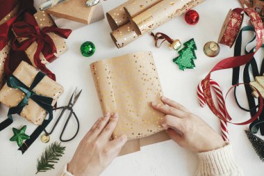 Mutlu Noeller! Şık Noel hediyelerini paketleyen eller dümdüz uzanıyordu. Modern hediye paketini hazırlayan kişi altın ambalaj kağıdı, kurdeleler, beyaz arka planda süsler. Atmosferik kış zamanı