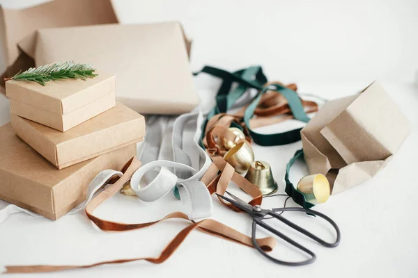 Wrapping stylish eco christmas gifts composition. Simple craft gift boxes, velvet ribbons, christmas bells, fir branch and scissors on white background. Merry Christmas! Sustainable holidays