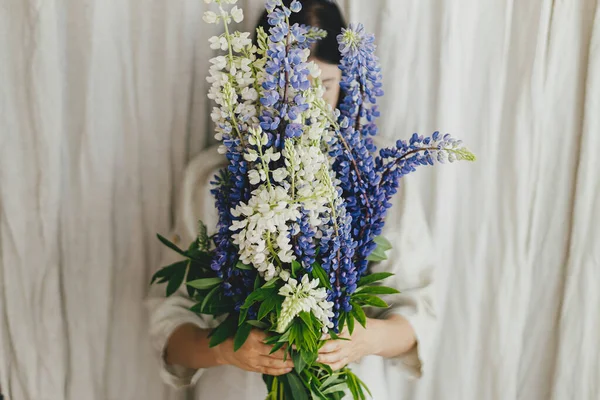Stylish woman behind lupine bouquet in rustic room. Gathering and arranging summer wildflowers at home in countryside. Lupine flowers close up in hands of young female in linen dress