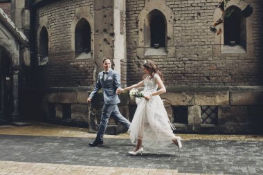 Beautiful emotional wedding couple running and smiling in european city. Provence wedding. Stylish happy bride and groom walking on background of church in sunny street