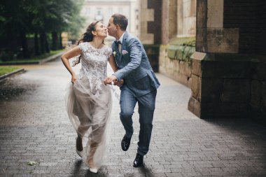 Beautiful emotional wedding couple smiling and kissing in rain in european city. Provence wedding. Stylish happy bride and groom running on background of old church in rainy street.