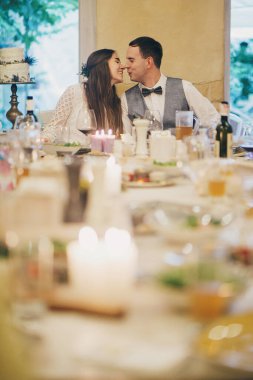 Beautiful wedding couple celebrating wedding on background of delicious festive table with food and drinks in restaurant. Stylish happy bride and groom kissing. Provence wedding reception