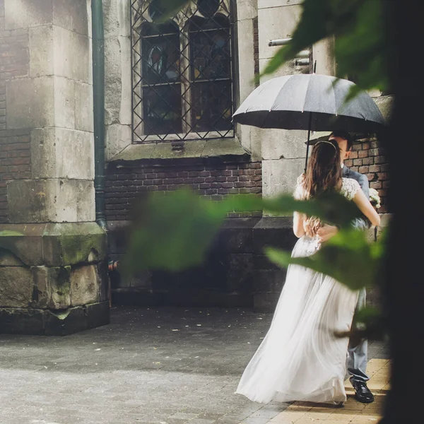 Stylish bride and groom kissing under umbrella on background of old church in rain. Provence wedding. Beautiful wedding couple embracing under black umbrella in rainy street. Romantic moment