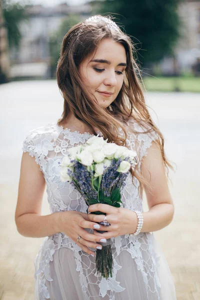 Beautiful bride in stylish vintage dress posing in sunny street of european city. Sensual bride in stylish gown walking with wedding bouquet of roses and lavender. Provence wedding