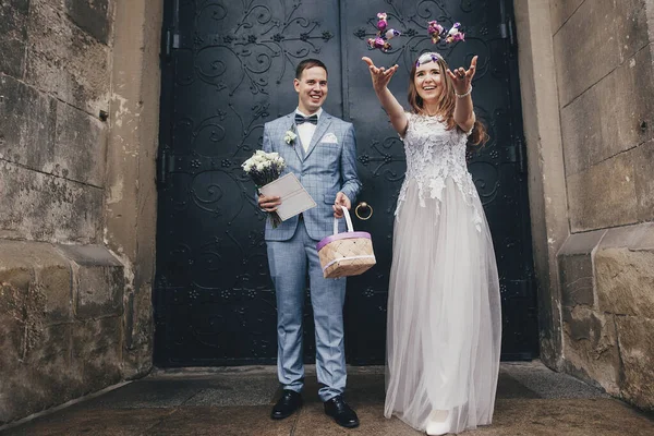 聖なる結婚後 スタイリッシュな幸せな花嫁と新郎は教会の背景にキャンディーを投げます 大聖堂での結婚式 お菓子と美しい感情的な精神的な結婚式のカップル — ストック写真