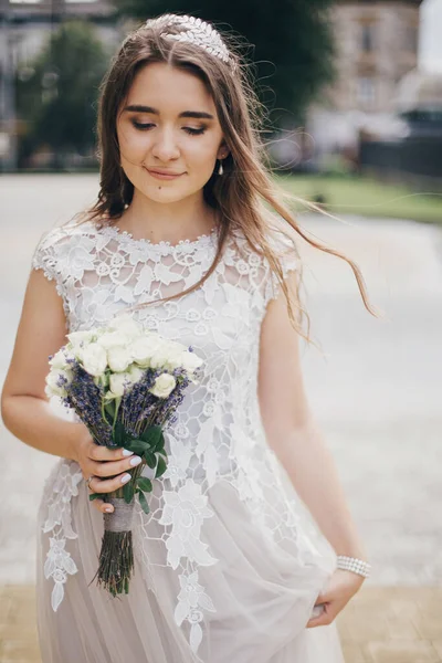 Beautiful bride in stylish vintage dress walking in sunny street of european city. Sensual bride in stylish gown walking with wedding bouquet of roses and lavender. Provence wedding
