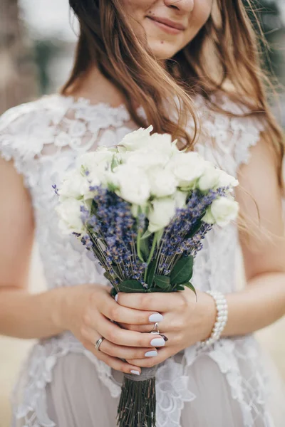 Portrait of sensual bride with wedding bouquet of roses and lavender in stylish gown. Provence wedding. Beautiful bride in stylish vintage dress posing in sunny street of european city