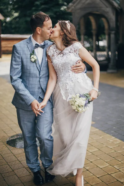 Beautiful emotional wedding couple kissing and walking in european city. Provence wedding. Stylish happy bride and groom walking on background of church in sunny street