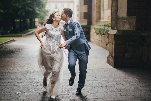 Beautiful emotional wedding couple smiling and kissing in rain in european city. Provence wedding. Stylish happy bride and groom running on background of old church in rainy street.
