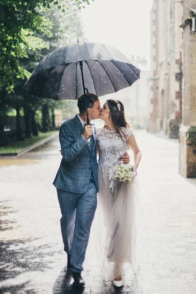 Stylish bride and groom walking under umbrella and kissing on background of old church in rain. Provence wedding. Beautiful wedding couple embracing together in sunny rainy street