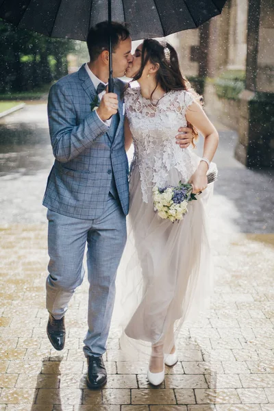 Stylish bride and groom walking under umbrella and kissing on background of old church in rain. Provence wedding. Beautiful wedding couple embracing together in sunny rainy street