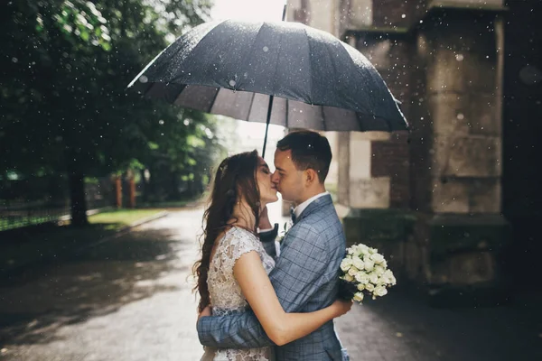 Blurred image of stylish bride and groom kissing under umbrella on background of old church in rain. Provence wedding. Beautiful wedding couple embracing under black umbrella in rainy street