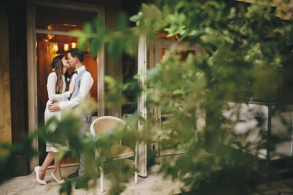 Stylish bride and groom gently embracing in stylish restaurant among trees. Beautiful romantic wedding couple kissing. Provence wedding reception