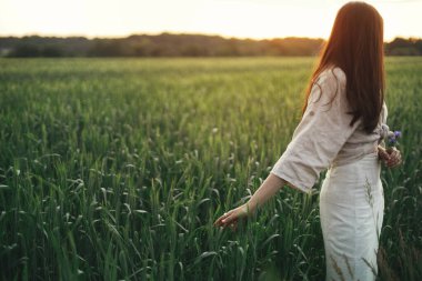 Woman relaxing in wheat field in warm sunset light. Stylish young female in rustic dress holding wildflowers in hands in evening summer countryside. Tranquil atmospheric moment