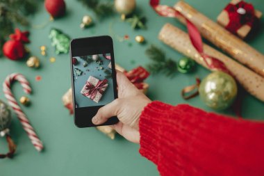 Şık bir şekilde paketlenmiş Noel hediyesi, altın kağıt, kırmızı kurdele ve yeşil arka planda şenlikli dekorasyonların fotoğrafını çekiyorlar. Sosyal medya. Mutlu Noeller ve mutlu tatiller!