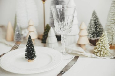 Modern Noel sofrası. Tabakta şık bir Noel ağacı, klasik çatal bıçak takımı, gözlük, şenlikli köknar ve beyaz kırsal masada evler. Masanın tatil düzenlemesi