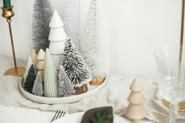 Noel Masası Dekorasyonu. Şık noel küçük ağaçları tabakta keten koşucusu, kırsal masada altın mumlar. Modern beyaz suni köknar ağacı süslemesi. Tatil düzenlemesi, masa ayarlama.