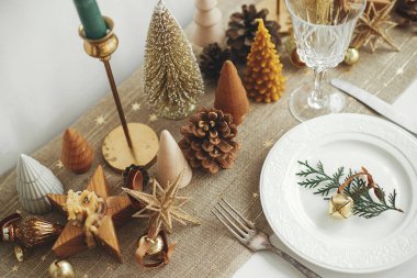 Şık bir Noel sofrası. Tabağında zil, kaliteli çatal bıçak, şarap kadehi, modern altın noel ağaçları ve kırsal masada mumlar olan fidan dalı. Tatil kahvaltısı, yeni yıl kutlaması.