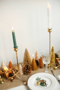 Şık bir Noel sofrası. Tabağında zil, kaliteli çatal bıçak, şarap kadehi, modern altın noel ağaçları ve kırsal masada mumlar olan fidan dalı. Tatil kahvaltısı, yeni yıl kutlaması.