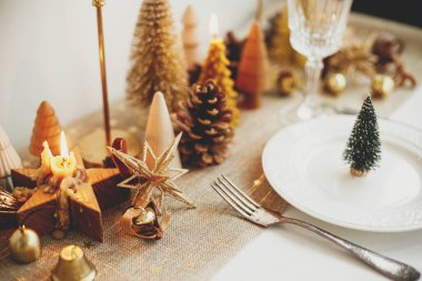 Şık bir Noel sofrası. Tabakta küçük bir Noel ağacı, klasik çatal bıçak takımı, şarap kadehi, modern altın noel ağaçları ve masada mumlar. Tatil kahvaltısı, yeni yıl kutlaması.