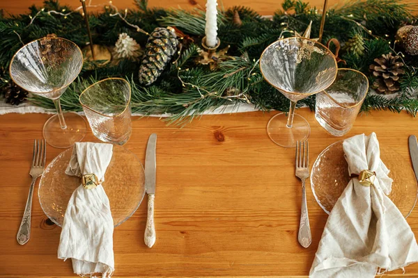 Christmas table setting. Linen napkin with bell on plate, vintage cutlery, glasses, fir branches with golden lights and pine cones on table. Holiday arrangement of table, top view