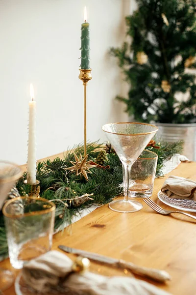 Christmas table setting. Linen napkin with bell on plate, vintage cutlery, glasses, fir branches with golden lights and pine cones on table. Holiday arrangement of table