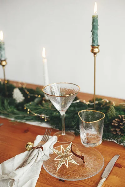 Stylish Christmas table setting. Linen napkin with bell, star on plate, vintage cutlery, wineglass, fir branches with golden lights, pine cones and candle on table.  Holiday brunch