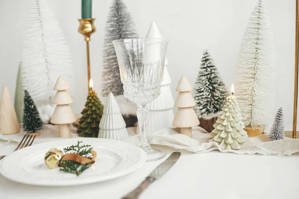 現代のクリスマステーブルの設定 プレート上の鐘と杉の枝 ヴィンテージのカトラリー お祝いの小さなクリスマスツリーや白素朴なテーブルの上の家 テーブルの休日の配置 — ストック写真