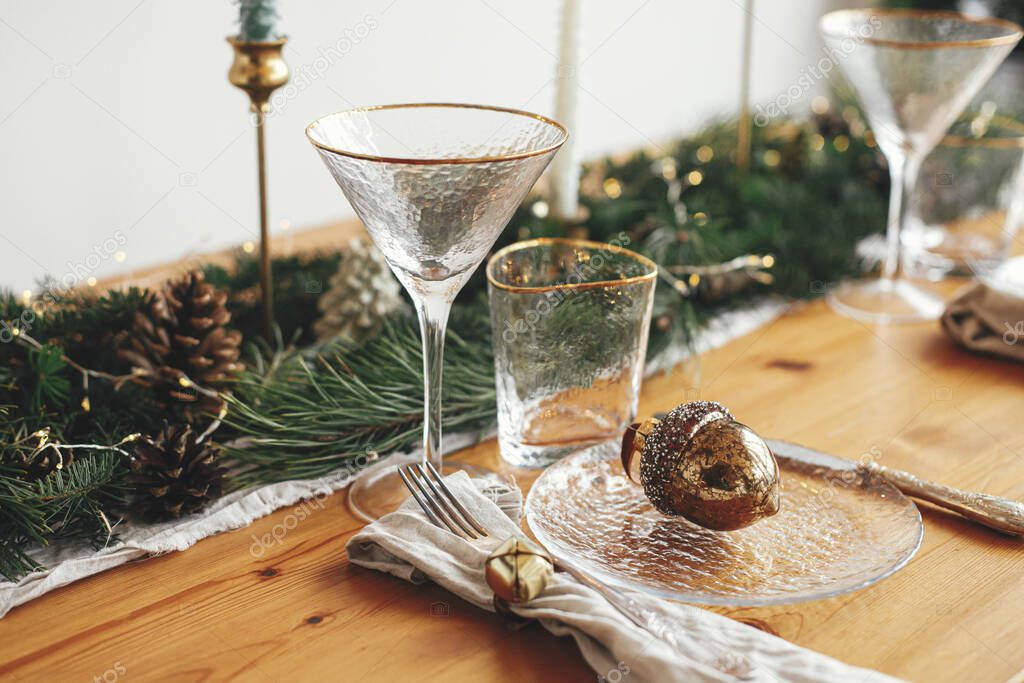 Christmas table setting. Linen napkin with bell, acorn on plate, vintage cutlery, glasses, fir branches with golden lights and pine cones on table. Holiday arrangement of table