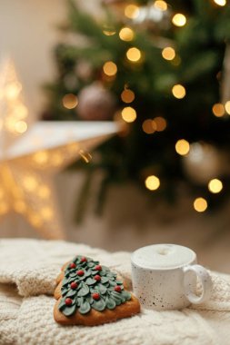 Sıcak bir fincan kahve ve yılbaşı ağacı kurabiyesi örülmüş süveterin üzerinde ışıklarla Noel ağacı. Atmosferik kış ikilisi. Noel afişi. Sıcak tatiller