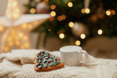 Atmosferik kış ikilisi. Sıcak bir fincan kahve ve yılbaşı ağacı kurabiyesi örülmüş süveterin üzerinde ışıklarla Noel ağacı. Noel afişi. Sıcak tatiller