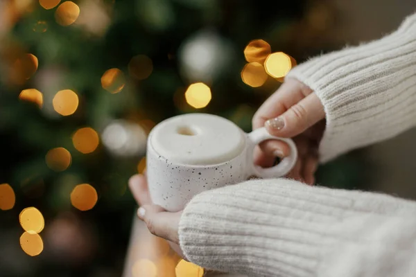 Hands holding warm cup of coffee on background of christmas tree with lights. Cozy home, atmospheric winter hygge. Woman hands in warm sweater holding  stylish mug at illumination bokeh