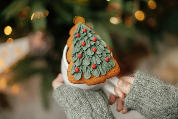 Merry Christmas! Holiday banner. Hands holding cup with christmas tree gingerbread cookie on background of christmas tree with lights. Cozy home, atmospheric winter time