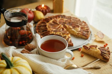 Atmosferik sonbahar natürmort. Tarçınlı, mumlu, sonbahar yapraklı, balkabaklı ve fındıklı taze elmalı turta ile sade ahşap masada, şık bir fincanda ılık çay. Hygge evde, rahat bir an.
