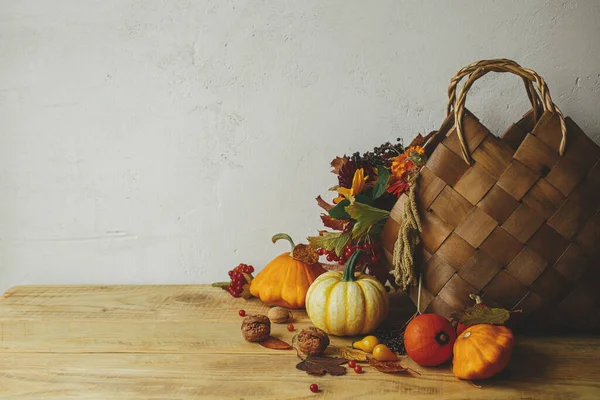 Autumn flowers in stylish basket, pumpkins, berries and nuts on rustic wooden table in room. Seasons greeting card. Happy Thanksgiving! Harvesting at farm. Atmospheric autumn still life