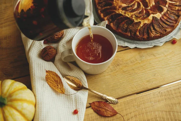 Cozy autumn. Pouring herbal tea in stylish cup on background of wooden table with freshly baked warm apple pie with cinnamon, autumn leaves, pumpkins and nuts. Hygge home